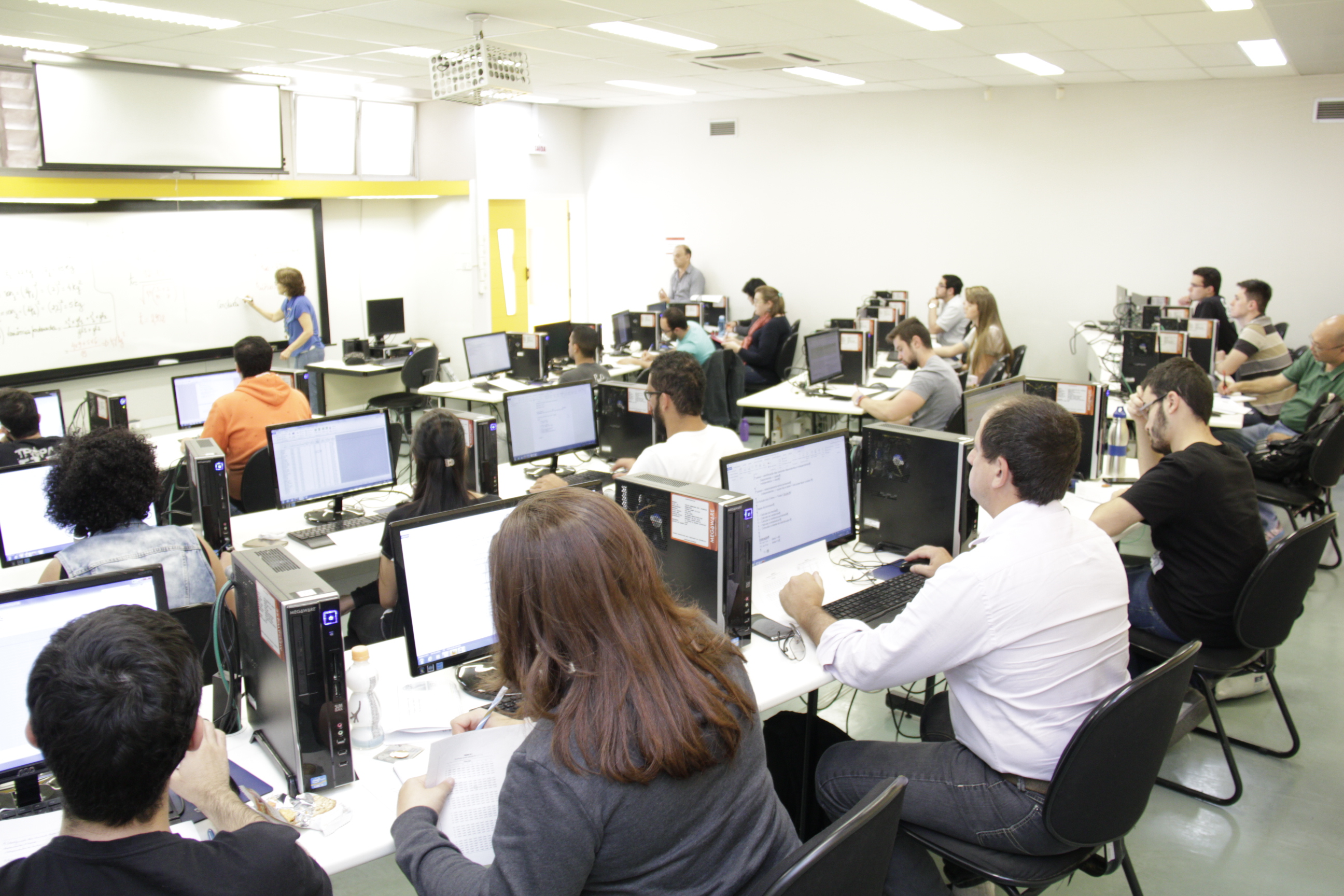 sala de aula com computadores pretos e alunos virados para lousa